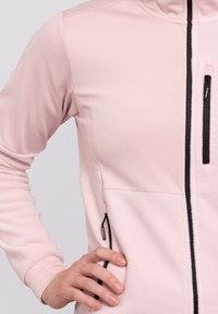 Veste rose clair au design ajusté. Présente des zips noirs et un matériau en polaire texturé sur la partie inférieure. Idéale pour le superposition.