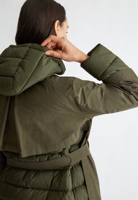 Olijfgroene pufferjas met een afneembare capuchon, gevoerd textuur, ceintuur in de taille en een grote kraag. Glad buitenmateriaal.