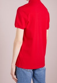 Polo rouge à manches courtes, fabriqué en tissu de coton. Doté d'un col classique et d'une coupe droite. Porté avec un jean denim bleu.