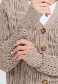 Beige knitted cardigan with a textured pattern, ribbed long sleeves, and three large brown buttons at the front.
