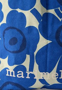 Μπλε ύφασμα με μεγάλα λουλουδάτα σχέδια σε σκούρο μπλε και ανοιχτό μπεζ, με τη λέξη «marimekko» σε λευκά γράμματα, τοποθετημένη στο κάτω μέρος.