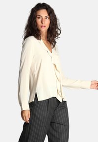 Blusa color crema con un davanti arricciato, maniche lunghe e una vestibilità morbida, abbinata a pantaloni a coste scure. Tessuto dalla consistenza liscia.