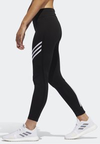 Mustat urheilulegginsit korkealla vyötäröllä, joissa on kolme valkoista raitaa sivussa, yhdistettynä valkoisiin lenkkareihin. Liukas, venyvä materiaali.