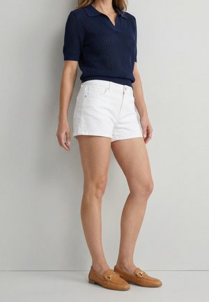 Kobieta ubrana w granatową dzianinową koszulkę polo z krótkim rękawem, białe szorty dżinsowe i beżowe skórzane mokasyny, stojąca na tle jednolitej jasnoszarej ściany.