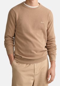 Pull beige en maille avec un motif texturé, col rond, poignets et ourlet côtelés, avec un petit logo blanc sur la poitrine. Associé à un pantalon beige.