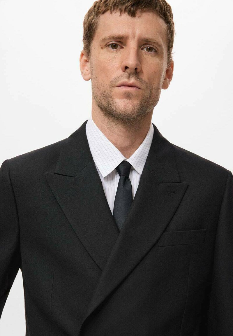 Schwarzer Anzug mit klassischem Schnitt und breiten Revers, kombiniert mit einem weiß gestreiften Hemd und einer schwarzen Krawatte, die eine glatte Textur zeigt.