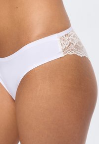 Λευκά εσώρουχα χωρίς ραφές με λεπτομέρεια δαντέλας στο ισχίο, φορεμένα από άτομο με μεσαίο καφέ δέρμα, εμφανιζόμενα από το πλάι της μέσης έως τον άνω μηρό.