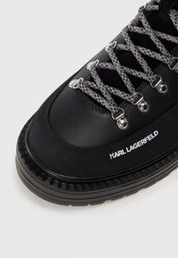 Zapatilla negra de cuero con ojales metálicos plateados, cordones grises y detalle de marca que dice "KARL LAGERFELD" en el lateral. Suela texturizada.