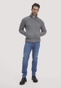 Grauer, gestrickter Reißverschluss-Pullover mit hohem Kragen, kombiniert mit blauen Jeans und schwarzen Schuhen. Der Pullover hat ein strukturiertes Muster.