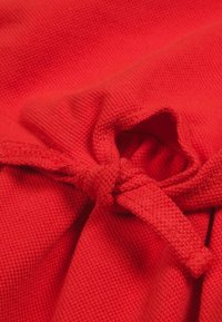 Czerwona tkanina o teksturowanym splocie, związaną kokardą na środku. Materiał jest gładki z lekkim rozciągnięciem.