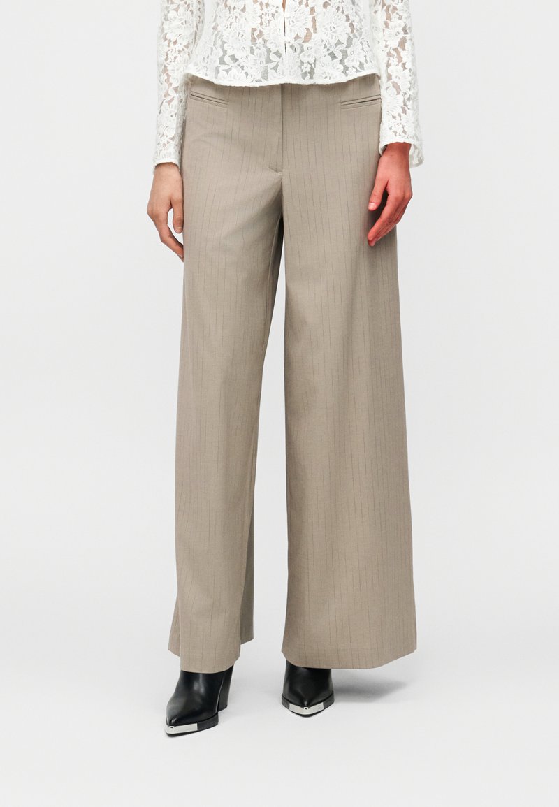 Γυναίκα με μπεζ παντελόνι φαρδιάς  γραμμής με ρίγες, λευκό μακρυμάνικο τοπ από δαντέλα και μαύρα μυτερά μποτάκια με τακούνι και μεταλλικές μύτες.