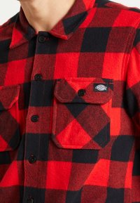 Camisa de franela a cuadros rojos y negros con dos bolsillos en el pecho, botones negros y una etiqueta en la solapa del bolsillo izquierdo. Textura suave.