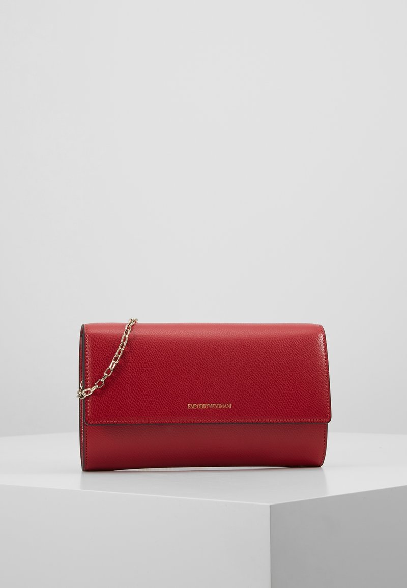 Pochette en cuir rouge Emporio Armani avec bandoulière chaîne dorée et logo doré sur le devant, posée sur un piédestal blanc contre un fond gris.