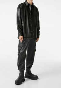 Camisa de piel sintética negra con botones, combinada con pantalones cargo a juego que tienen bolsillos laterales, y botas negras de tobillo gruesas.