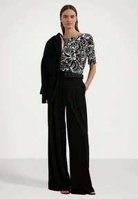 Sieviete, tērpta melnās platās biksēs, melnā un baltā ziedu topā, tur melnu jaku uz pleca, stāv pret vienkāršu fona.