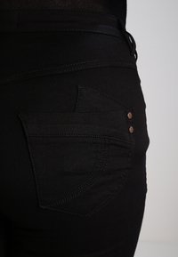 Jeans de mezclilla negra con un diseño de bolsillo trasero curvado, botones de cobre en contraste y costuras tonales sutiles. Textura suave, forma ajustada.