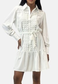 La robe-chemise blanche présente un devant boutonné avec de la dentelle décorative, des manches longues, une ceinture nouée à la taille et une jupe évasée avec un ourlet à volants.