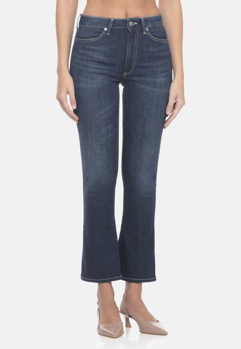 Jeans a campana di colore blu scuro realizzati in denim con cinque tasche, cuciture a contrasto e chiusura con zip. Sono abbinati a scarpe con tacco nel tono nude.