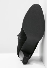 Botte de cheville en daim noir avec un talon texturé, détail de fermeture éclair sur le côté et semelle en caoutchouc lisse. La botte est inclinée contre un fond blanc.