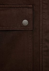 Échantillon de tissu brun avec une surface texturée. Présente une poche plaquée, des bords cousus, et une étiquette métallique circulaire avec la marque.