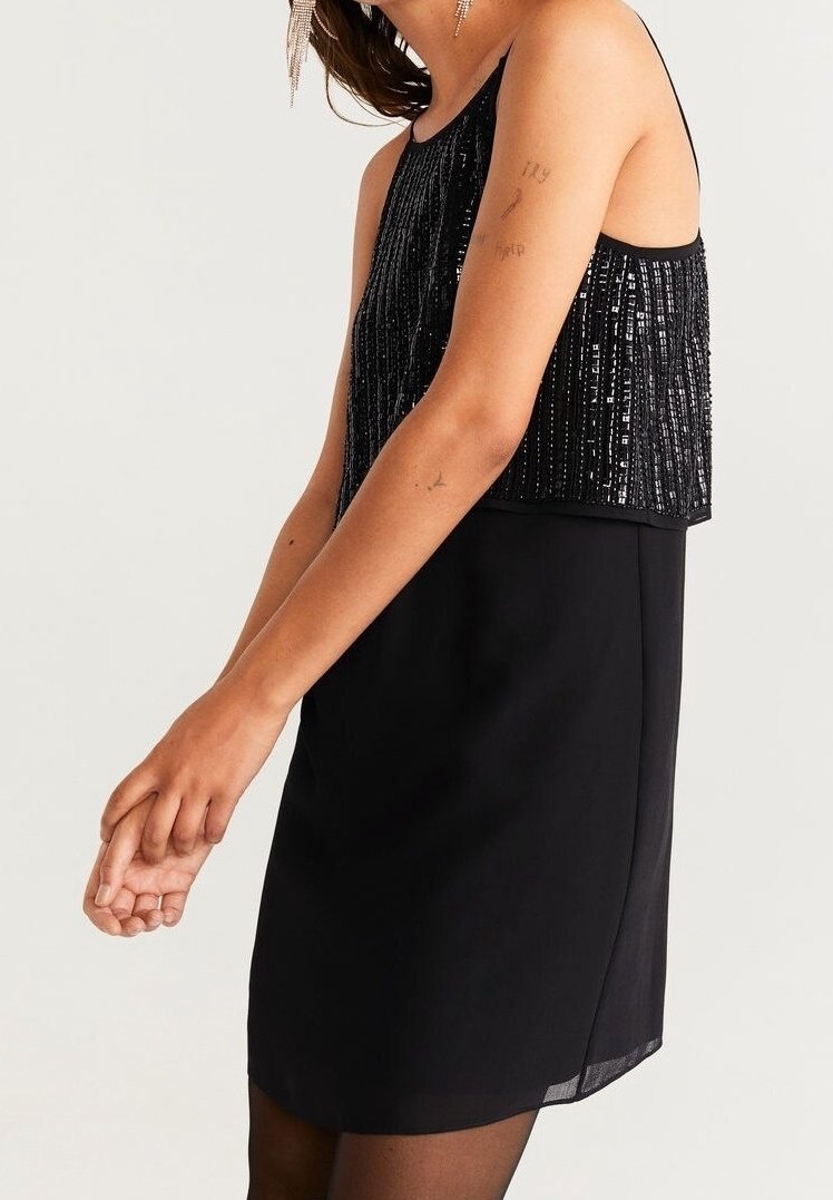 Femme portant une robe noire sans manches avec un haut pailleté, debout les mains jointes devant un fond clair.