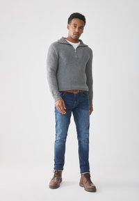 Šedý ribbed pulovr s límcem na půl zip, nošený přes bílou košili. V kombinaci s modrými džíny a hnědými šněrovacími botami. Jednoduchý design, bez vzorů.