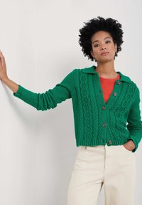 Grön stickad kofta med ett texturerat mönster, krage och bruna knappar, kombinerad med en röd topp och krämfärgade byxor.