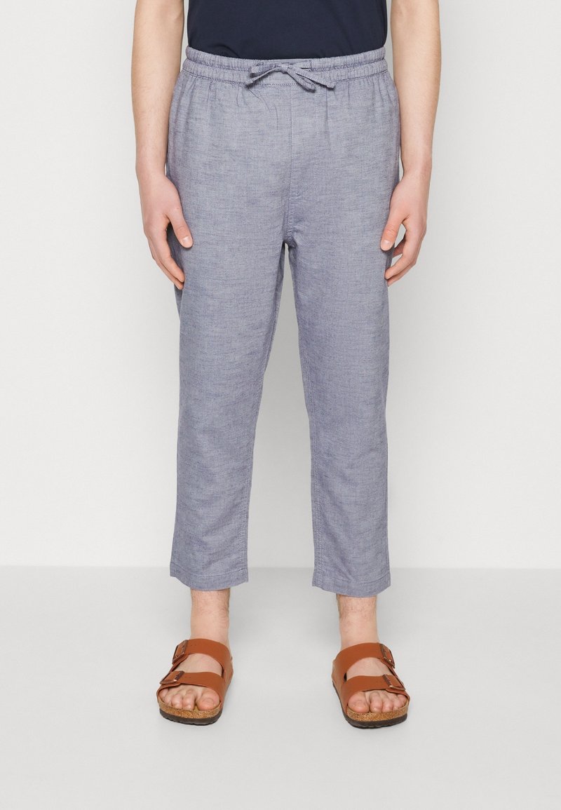 Pantalon gris clair en mélange de lin avec une taille élastique et un cordon de serrage, présentant une coupe décontractée et une longueur raccourcie, associé à des sandales marron.