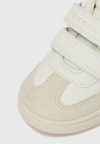 Baskets blanches et beiges avec des straps en velcro. Elles présentent une tige en cuir lisse et un bout en suède texturé. Logo affiché sur la sangle.