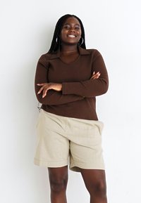 Top de manga larga marrón con cuello, combinado con pantalones cortos de lino beige claro. El modelo tiene el cabello trenzado y lleva pendientes en forma de aro.