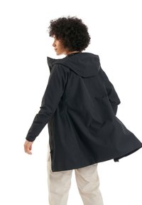 Schwarze wasserdichte Jacke mit Kapuze, mit lockerer Passform, Reißverschluss vorne und Seitentaschen, getragen über hellfarbigen Hosen.