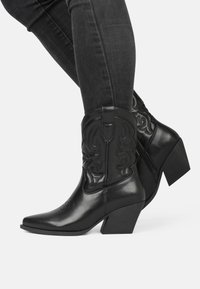 Botas de cowboy de cuero negro con punta afilada, tacón cuadrado y costuras decorativas. Cuentan con un zipper en el costado para facilitar su uso.