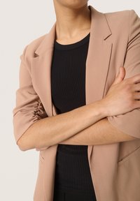 Béžový blazer s texturovanou tkaninou, zkrácenými rukávy a zapínáním na jeden knoflík, nošený přes černý žebrovaný top. Ruce jsou zkřížené.