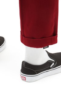Scarpe Vans nere slip-on con suole bianche, abbinate a calzini bianchi fino alla caviglia e pantaloni rossi arrotolati, con un'etichetta Vans sul polsino.