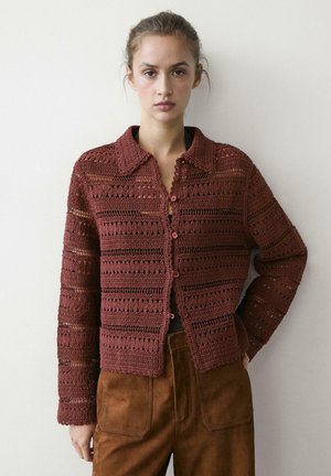 Giovane donna che indossa una giacca all'uncinetto color marrone rossiccio con bottoni e pantaloni in suede marroni, in piedi davanti a uno sfondo chiaro e semplice.