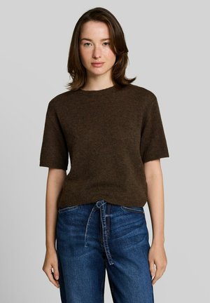 Giovane donna con capelli fino alle spalle che indossa un maglione marrone a maniche corte e jeans blu con cintura annodata in vita, in piedi davanti a uno sfondo grigio uniforme.