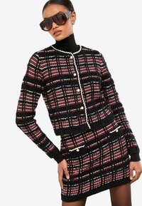 Černý a červený kostkovaný tvídový kabát s krátkým střihem, perlovými knoflíky a laděnou sukní. Má žebrované manžety a texturovaný vzor.