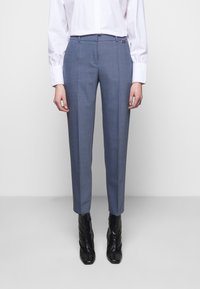 Pantalon bleu sur mesure en tissu texturé, coupe slim, avec poches latérales, associé à une chemise blanche à boutons et des bottines noires.