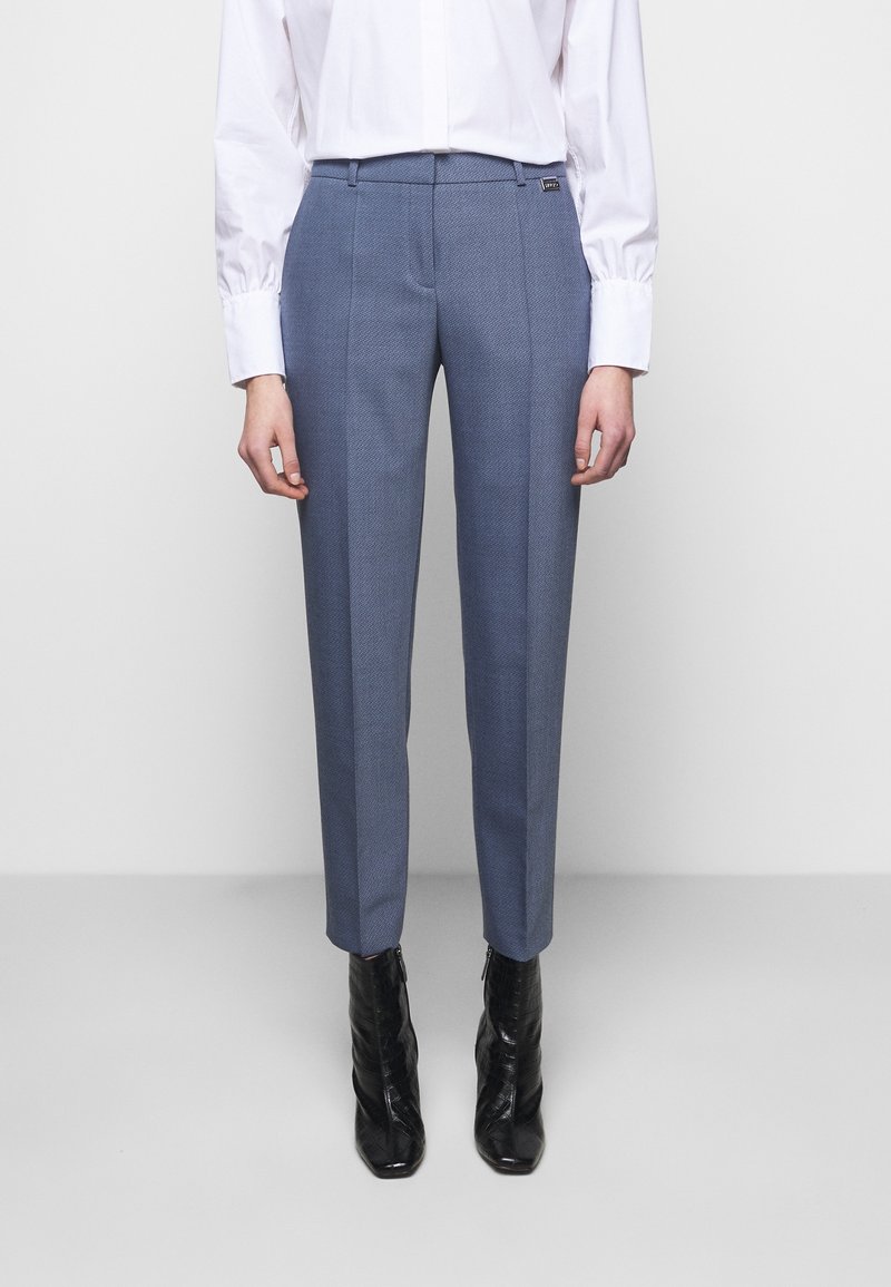 Pantalon bleu sur mesure en tissu texturé, coupe slim, avec poches latérales, associé à une chemise blanche à boutons et des bottines noires.