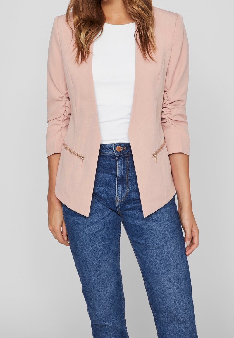 Femme portant un blazer rose clair avec des manches froncées, un haut blanc et un jean bleu taille haute sur un fond gris uni.