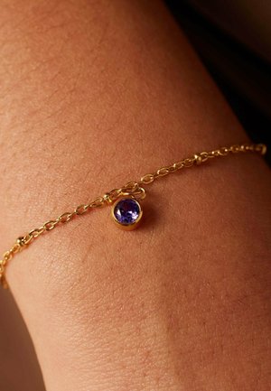 Bracelet en or avec une petite médaille ronde contenant une gemme violette. La chaîne présente un design lié intriqué, reposant sur la peau.