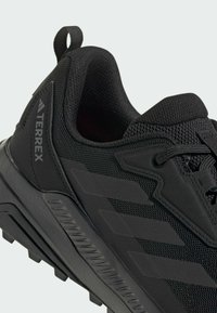 Chaussure de sport noire avec tige en mesh et semelle en caoutchouc. Présente un design texturé et trois bandes diagonales sur le côté. Logo de la marque visible.