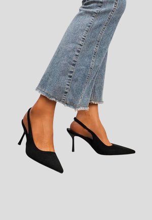 Personne portant un jean bleu raccourci et des escarpins noirs à bout pointu avec bride arrière, jambes légèrement croisées sur un fond uni.