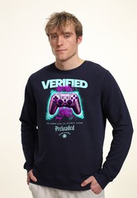 Felpa navy con grafica di un controller di gioco viola e testo "VERIFIED GAMER" in turchese e rosa. Tessuto morbido, design con colletto a girocollo.