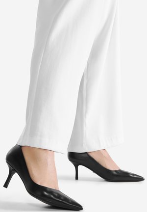 Zapatos de tacón alto de cuero negro con punta puntiaguda y tacón de aguja delgado, combinados con pantalones anchos blancos. Textura suave, diseño minimalista.