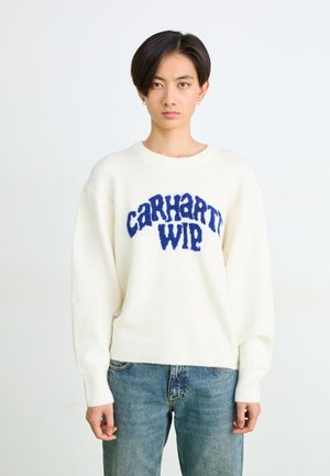 Pull en tricot blanc avec un logo "Carhartt WIP" bleu duveteux. Il a un col rond et des poignets côtelés, associé à un jean en denim bleu.