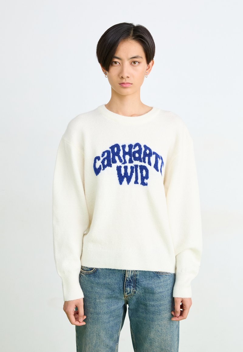 Witte gebreide trui met een zachte blauwe "Carhartt WIP"-logo. Voorzien van een ronde halslijn en geribbelde mouwen, gecombineerd met blauwe spijkerbroek.