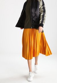 Sort satin bomberjakke med olivengrønne ærmer, kombineret med en lys gul plisseret midinederdel og hvide sneakers.