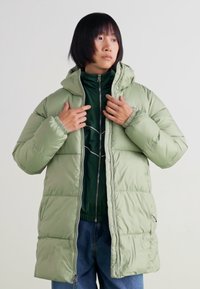 Jachetă puf verde cu glugă, cu un design matlasat și închidere cu fermoar, peste un strat interior verde închis și blugi.