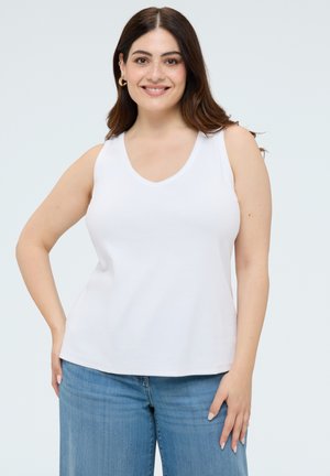 Lachende vrouw met lang bruin haar, gekleed in een wit mouwloos topje en blauwe spijkerbroek, staand met één hand op haar heup.